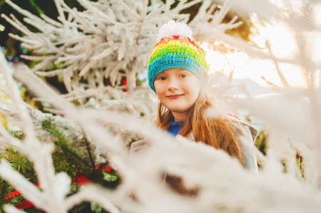 Sweet little girl wearing colorful hat, standing between christmas trees at sunsetの写真素材