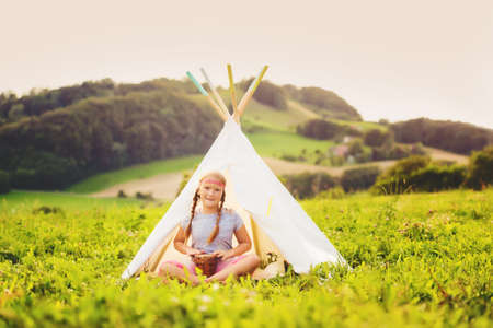 Cute little girl having fun outdoors, playing with tambour, sitting next to teepeeの写真素材