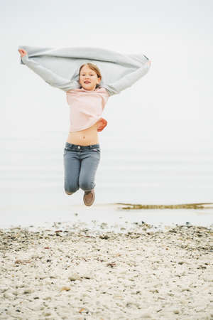 Adorable little girl of 8-9 years old playing by the lake, wearing grey trousers and long cardigan, jumping in the airの写真素材