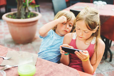 Two kids playing electronic games on the phone while waiting food orders in restaurantの写真素材