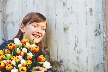 Portrait of cute 8-9 year old girl, holding bright bouquet of colorful fresh tulips, standing against white wooden background, laughing kid, eyes closedの写真素材