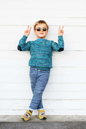 Fashion kid in sunglasses, wearing blue knitted pullover, joggers and yellow basket shoes, standing against white background, showing peace signの写真素材