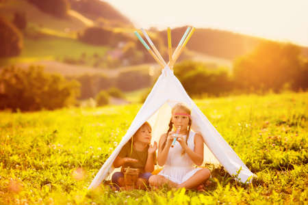 Two happy kids sitting in a tent on a nice warm evening, playing flute and drum music. Brother and sister having good time togetherの写真素材