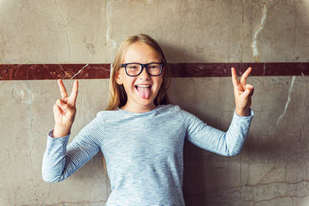Interior portrait of a cute school girl wearing glasses, stripy shirt, showing peace sign, pulling a tongueの写真素材