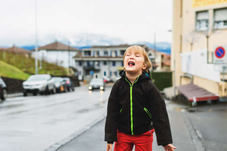 Little boy playing outdoors in early spring, walking next to road, wearing black waterproof jacketの写真素材
