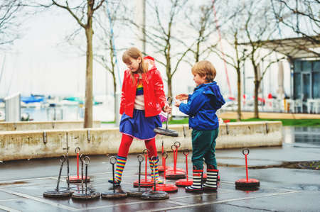 Group of two kids playing giant checkers on playground, wearing rain coats and boots. Image taken in...