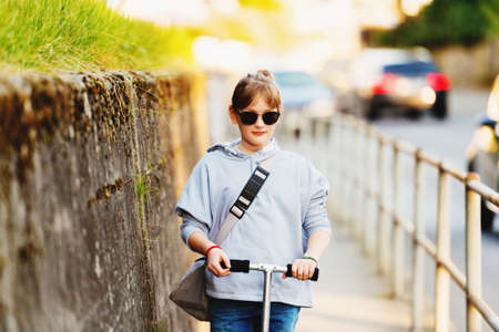Happy 9 year old school girl with backpack in the streets of the city, riding scooter, wearing sunglassesの写真素材