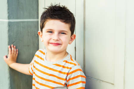 Outdoor portrait of a cute little boy standing against grey wall on a very sunny day, wearing grey and orange stripe tshirtの写真素材