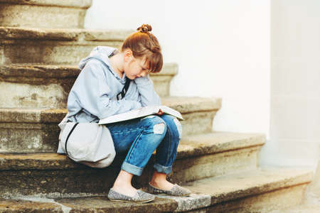 Kid girl reading book outside, sitting on stairs, wearing grey sweatshirt and backpackの写真素材