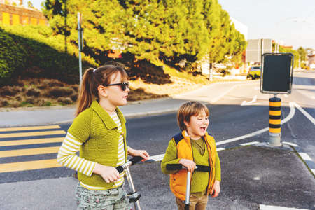 Two happy kids playing outdoors, fashion boy and girl posing on the street, riding scooters, crossing the roadの写真素材