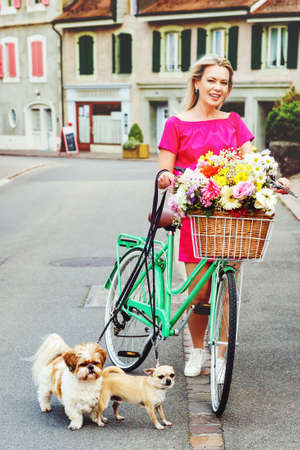 Beautiful blond woman wearing bright pink dress, riding a bicycle with big colorful bouquet of flowers in basket and holding leashes with two small dogs. Image taken in Lausanne, Switzerlandの写真素材