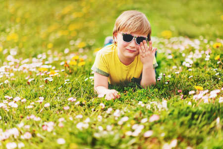 Sunny summer portrait of young handsome little boy playing outdoors on a nice warm day, lying on bright green grass, wearing yellow t-shirt and sunglassesの写真素材