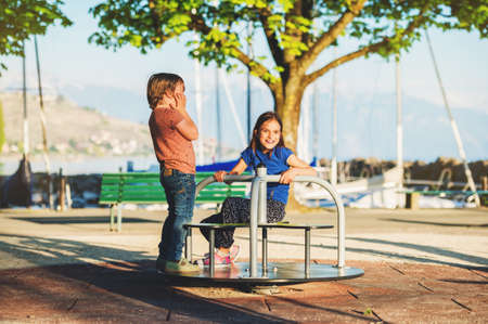 Two kids having fun on playground. Stylish kids playing on merry-go-round in the park on a very sunny day. Adorable children friends spending time togetherの写真素材