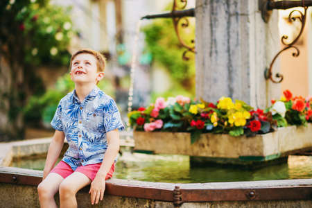 Outdoor summer portrait of adorable kid boy resting outdoors next to fountain on a very hot sunny day, wearing blue print shirt and red shortsの写真素材
