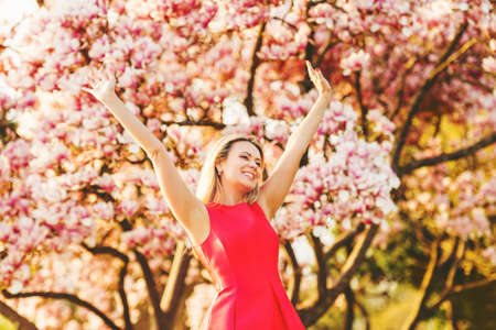 Beautiful happy young woman enjoying warm sunny day in spring garden, wearing bright red dress, arms wide openの写真素材