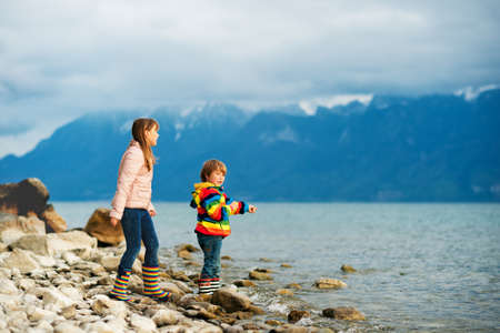 Two happy kids, little brother and big sister, playing together by lake Geneva on a very cloudy day with swiss mountains Alps on background. Image taken in Lausanne, Switzerlandの写真素材