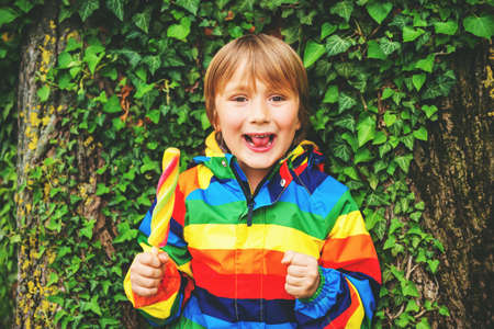 Cute little boy eating ice cream outdoors, wearing colorful ice cream, leaning on the treeの写真素材