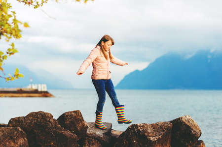 Adorable kid girl playing by the lake on a very cloudy day, wearing warm jacket and rain bootsの写真素材