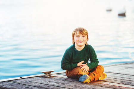 Outdoor portrait of adorable 5-6 year old boy resting by the lake, wearing green pullover and yellow trousersの写真素材