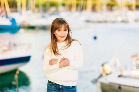 Outdoor portrait of pretty little girl posing by the lake, wearing warm white pullover, arms crossedの写真素材