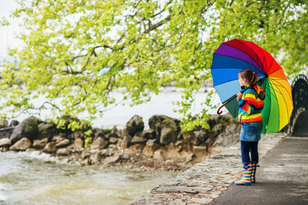 Outdoor portrait of pretty little kid girl with big colorful umbrella, wearing rain jacket and boots, weather concept, fashion for kidsの写真素材