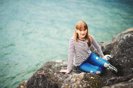 Adorable kid girl resting by the lake, wearing stripe t-shirt and fashionable ripped jeansの写真素材