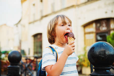 Cute little boy eating chocolate ice cream outdoors, wearing backpack, travel with kidsの写真素材