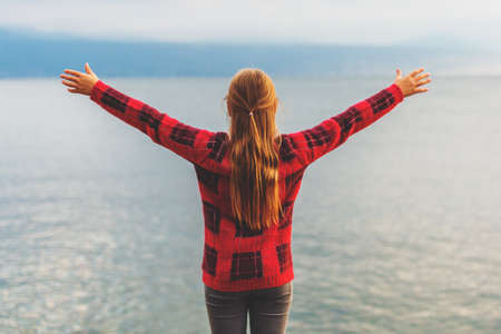 Kid girl enjoying nice autumn day ba the lake; wearing warm red pullover, arms up wide open, back view.の写真素材