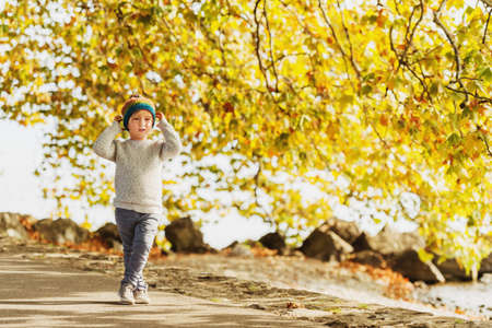 Little boy playing by the lake on a sunny day, wearing warm hat, grey knitted pullover, trousers and shoesの写真素材