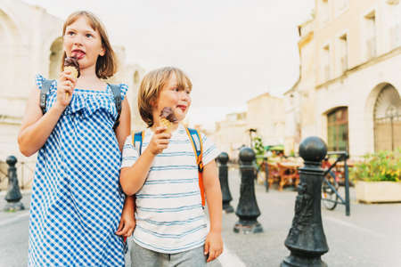 Two funny kids tourist on the street of old european city, children enjoying summer vacation on south of France. Little boy and girl eating chocolate ice cream, wearing backpacksの写真素材