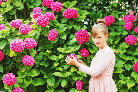 Outdoor portrait of pretty little girl playing with beautiful hydrangea flowers in summer garden, wearing ballerina style dress, hair bun and diamond tiaraの写真素材