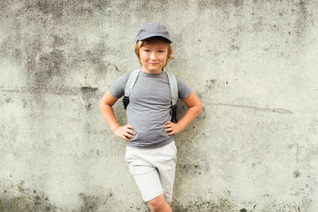Outdoor portrait of funny little boy standing next to grey wall, wearing cap and backpack