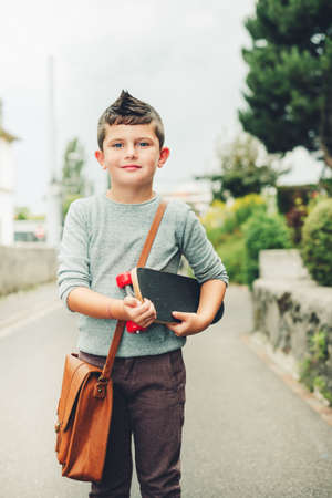 Outdoor portrait of funny little schoolboy wearing brown leather bag over shoulder, holding skateboard. Back to school concept. Film look toned imageの写真素材
