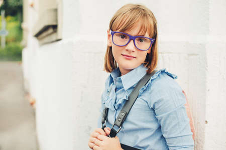 Outdoor portrait of a cute little 9-10 year old girl wearing blue uniform dress, backpack and glasses. Back to school conceptの写真素材