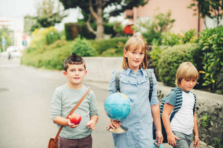 Group of three funny kids wearing backpacks walking back to school. Girl and boys enjoying school activities. Globe, lunch box, red apple and bag accessories.の写真素材