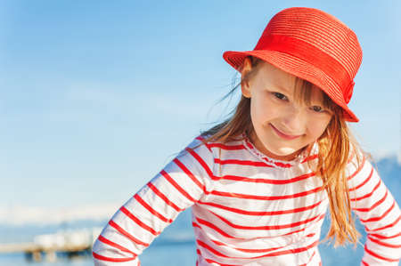 Outdoor portrait of adorable little kid girl against sunny blue sky, wearing red hat and stripe shirtの写真素材