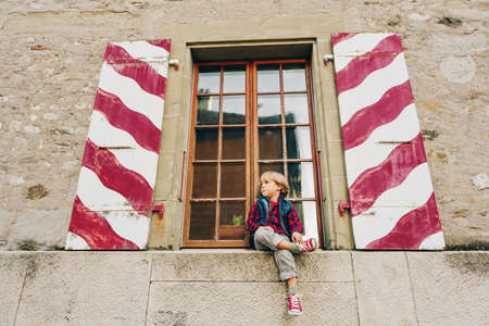 Outdoor portrait of funny little boy sitting o the window, wearing tennis shoes
