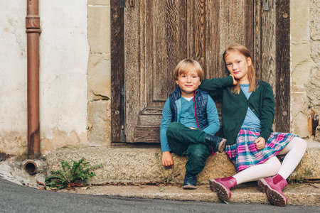 Group of two stylish kids posing outdoors, resting next to old vintage door. Fashion for schoolkids. Footwear and clothing for school. Little boy and girl playing togetherの写真素材