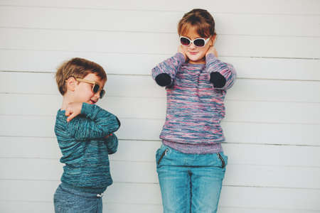 Group of two funny kids playing together outside, little boy and girl posing against white wooden background, brother and sister wearing matching pullovers, fashion for children, knitwear.の写真素材