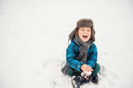 Cute little boy playing in winter park. Kid having fun outdoors, sitting on the snow, wearing warm blue jacket, hat and scarfの写真素材
