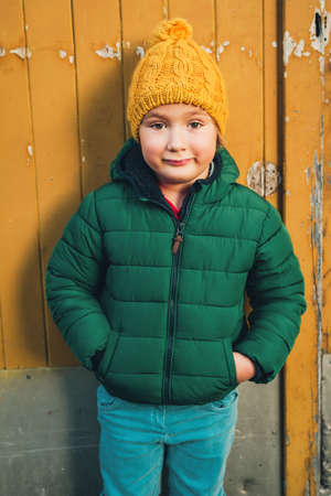 Outdoor portrait of a funny 4-5 year old little boy wearing green winter jacket, blue denim jeans,...