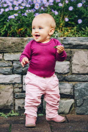 Outdoor portrait of adorable 9-12 month old baby girl playing with purple flowers, wearing pink...