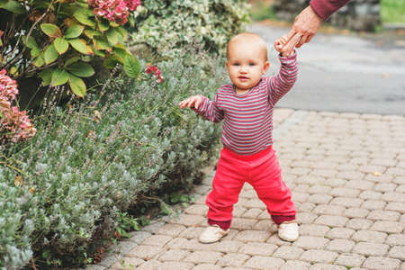 Adorable baby girl of 9-12 months old playing outside, wearing pink body and joggers, holding...
