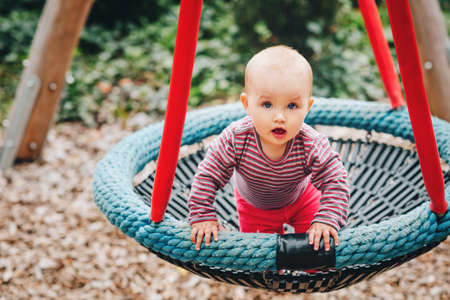 Baby girl having fun in the park, 9-12 months old kid playing in the big swing, summer playground,...