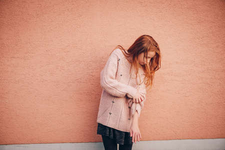 Outdoor portrait of adorable red-haired preteen girl wearing soft pink pullover, kid model posing against beige wall background. Fashion for young teensの写真素材