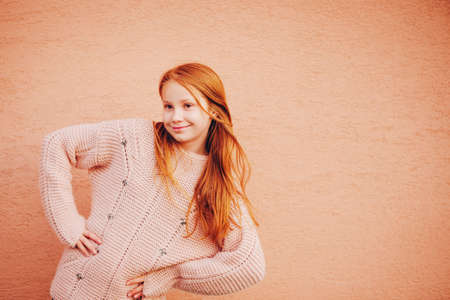 Outdoor portrait of adorable red-haired preteen girl wearing soft pink pullover, kid model posing against beige wall background. Fashion for young teensの写真素材