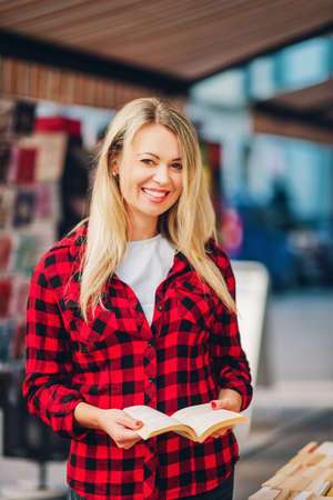 Happy young blond reading woman in a book store, wearing red plaid shirtの写真素材