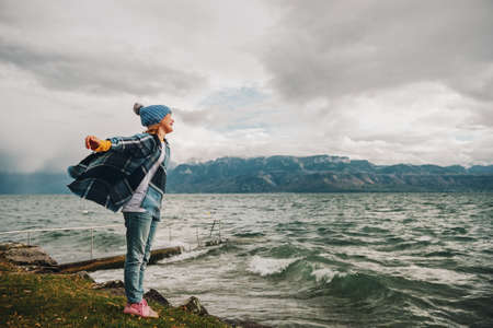 Sweet little girl playing by the lake on a very windy day, wearing blue hat and plaid poncho. Image...