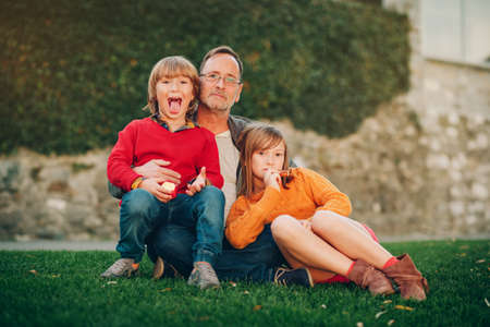 Outdoor portrait of happy father with two kids, boy and girl resting together in the park, relaxing on bright green grassの写真素材