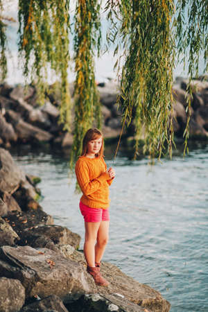 Outdoor portrait of cute preteen kid girl playing by the lake at sunset, wearing yellow pullover and red shirtsの写真素材